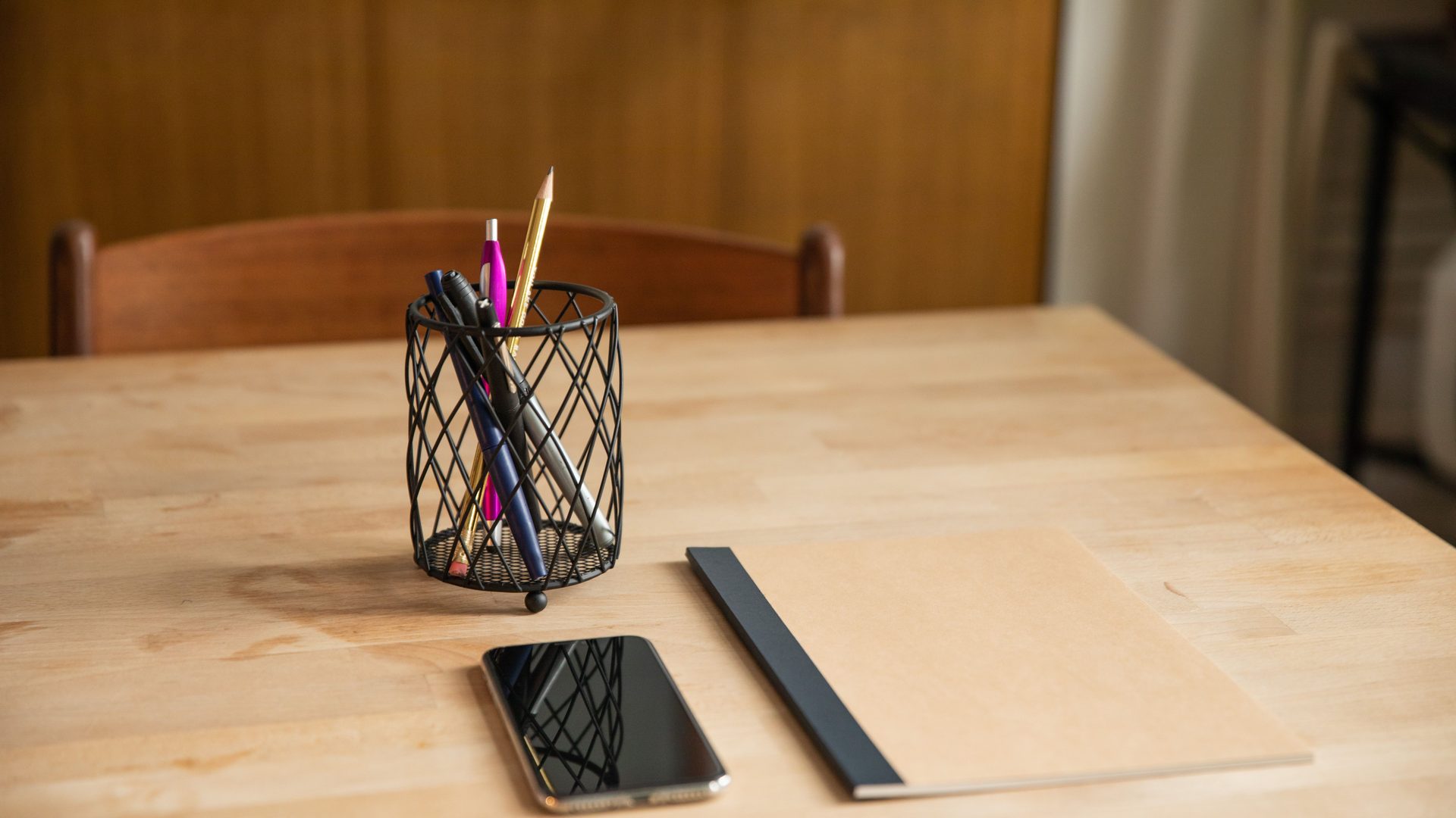 A jar of pens and pencils on a bare wooden table in daylight