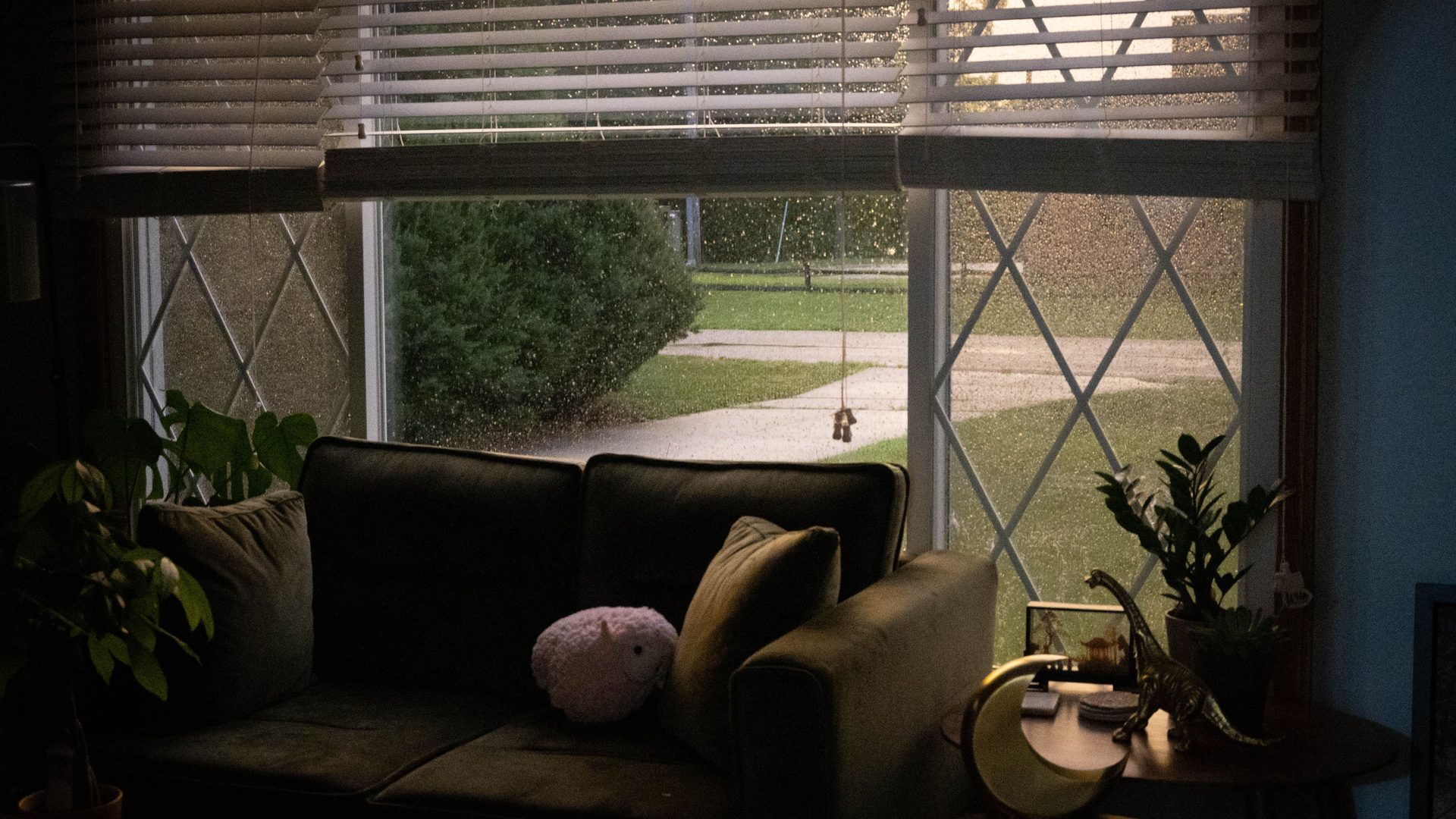 A sofa by a large window looking onto a driveway and lawn in the rain